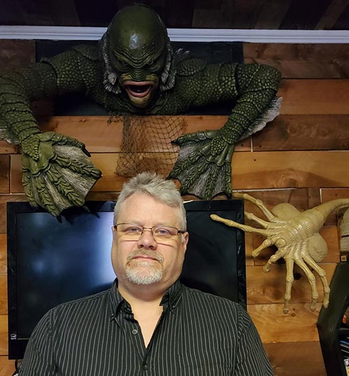 David Adkins in front of a TV, a green creature and a tan alien