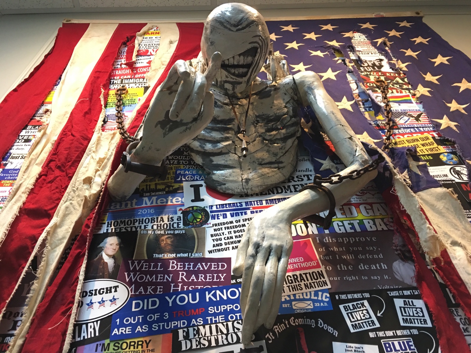 Torso, arms and hands of a chained body attached to a vertical wall with a tattered flag over various political prints