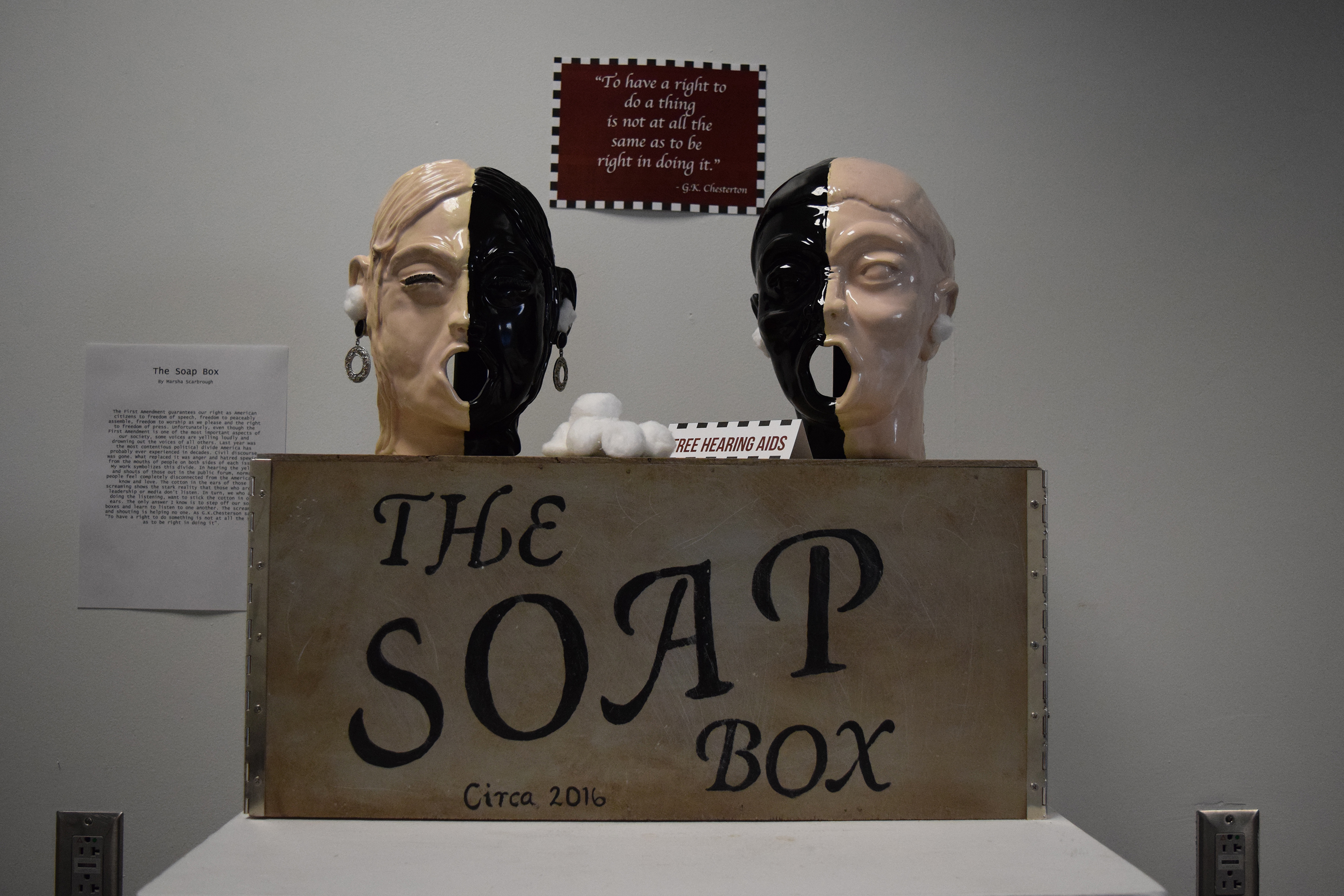 Box with "The Soap Box" written on the front side. On top of the box are two heads, each split with one side tan and the other black. Cotton balls are in the ears. One head has earrings.