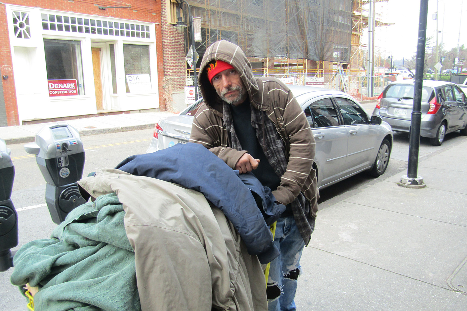 Photograph of a homeless man with a cart. The background is a city street with parked cars.