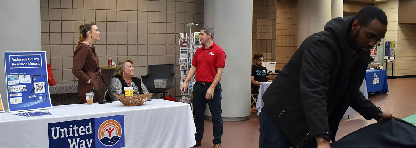 Individuals stand talking in front of a 'United Way' table. Several other individuals are around other tables.