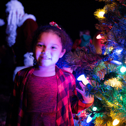 A child standing near a colorful Christmas tree with Santa in the background.