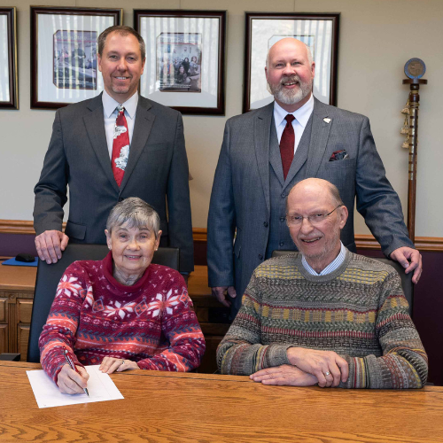 Two people sitting at a table, signing a piece of paper, with two others standing behind them.