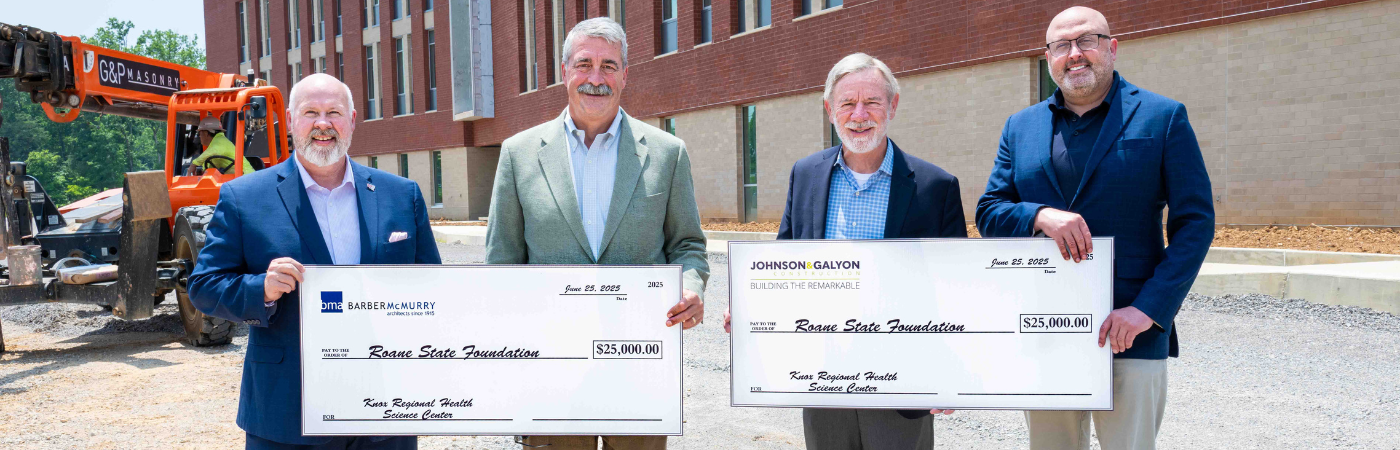 Four people standing at a construction site holding two large checks for $25,000 each.