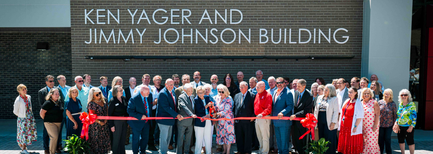 A large group of people in front of a brick building prepare to cut a red ribbon.