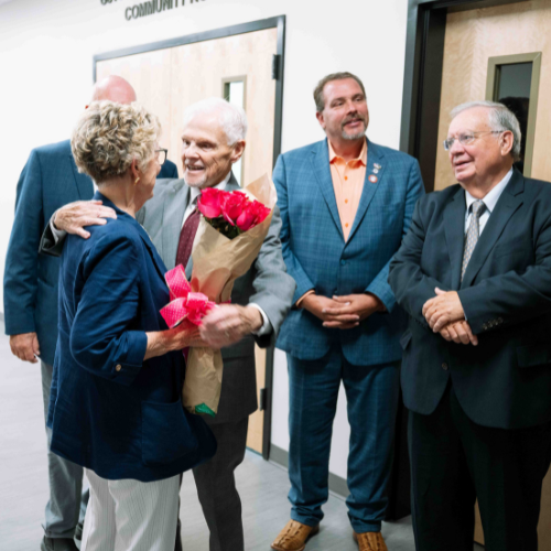 A woman holding a bouquet of flowers is congratulated by a group of people who are smiling.